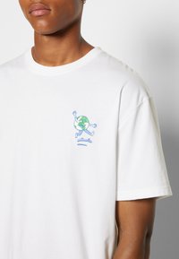 Lähellä oleva kuva henkilöstä, joka pukeutuu valkoiseen t-paitaan, jossa on pieni brodeerattu piirros Maapallosta, joka skeittaa vasemmassa rinnassa.