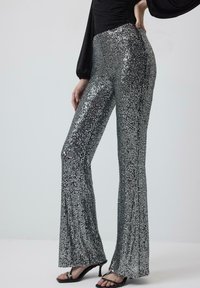 Zilveren pailletten flared broek met een gestructureerd oppervlak, voorzien van een hoge taille en een glanzende afwerking. Gepaard met zwarte hoge hakken sandalen.