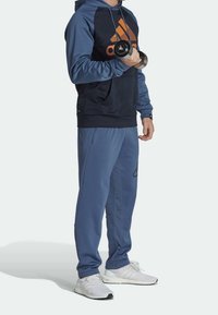 Navy and blue tracksuit with a large orange Adidas logo, textured sleeves, and black accents; holding a dumbbell. White athletic shoes.