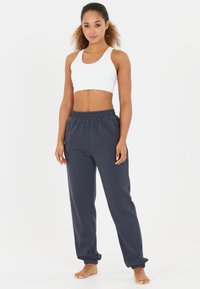 Weißer Sport-BH und dunkelgraue Jogginghose, aus Baumwolle, lockere Passform, elastischer Bund, umschlagbare Knöchel, barfuß sichtbar.