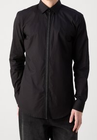 Camisa negra de botones, hecha de tela suave, con una cremallera central, mangas largas, cuello clásico y un dobladillo recto.