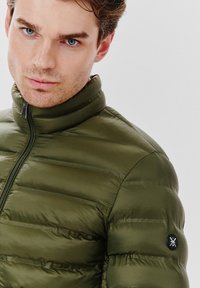 Wypełniona oliwkowa zielona kurtka z wysokim kołnierzem, z zamkiem błyskawicznym z przodu i okrągłą czarną naszywką z logo na lewym rękawie. Gładka tekstura.