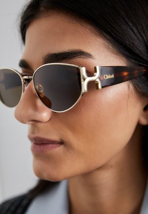 Gafas de sol redondas con un marco de metal dorado y lentes oscuros. Patillas de carey con el logo de Chloé. Presenta un acento de hardware distintivo en la parte frontal.