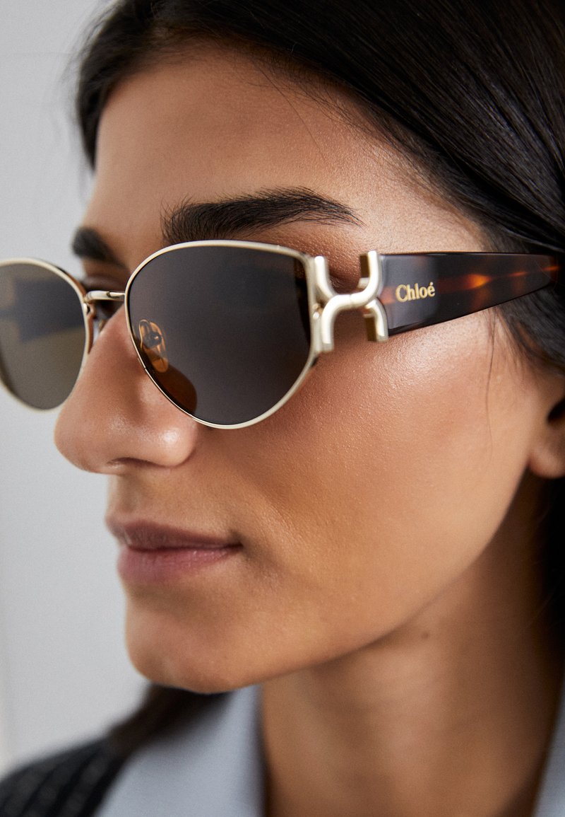Ümmargused prillid kullavärvi metallraami ja tumedate läätsega. Karikakra stiilis templid Chloé logoga. Esiküljel on silmapaistev metallist aksessuaar.