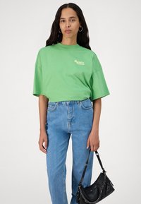 Camiseta verde de gran tamaño con mangas cortas y logo de "Pegador", combinada con jeans ajustados de cintura alta y un bolso negro de hombro.