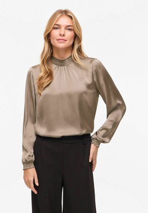 Blond kvinne som har på seg en taupefarget satengbluse med lange ermer, smocked hals og mansjetter, kombinert med høytaljede svarte bukser.