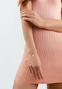 Pulsera de plata con un charm en forma de corazón y una superficie texturizada, llevada en una mano contra un vestido de rayas rosa.