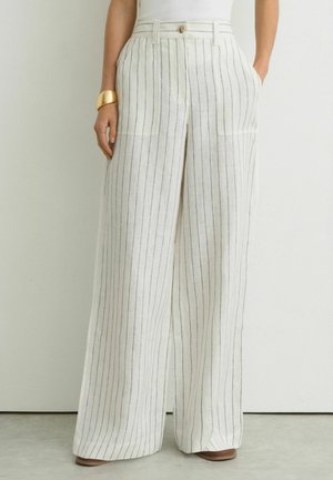 Naine, kes kannab kõrge vöökohaga laia säärega valgeid pükse, millel on õhukesed mustad vertikaalsed triibud, esimesed taskud ja nööpkinnitusega, sobitatud valge topiga.