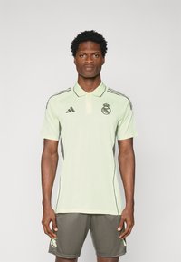 Lichtgroene sportpoloshirt met een kraag, donkergrijze zijaccenten en het Real Madrid-logo. Draagt een bijpassende donkergrijze short.