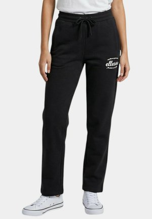 Pantalon de survêtement noir à cordon avec le logo blanc "ellesse" sur la cuisse gauche, porté avec des baskets blanches et un t-shirt blanc rentré.