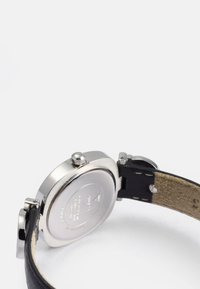 Roestvrijstalen horloge met een ronde kast, zwarte leren armband en een gestructureerde beige onderkant. Voorzien van een kroon en een doorzichtige achterkant.