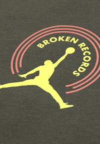 Σκούρο πράσινο ύφασμα με κίτρινη σιλουέτα ενός άλματος ανθρώπου με ένα μπάσκετ, περιτριγυρισμένη από ροζ κυκλικές γραμμές και το κείμενο "BROKEN RECORDS."