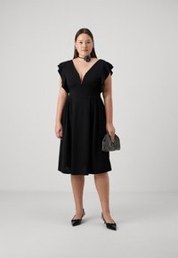 Vestido negro con mangas acampanadas, escote en V profundo, falda amplia y una textura ligera. Combinado con un bolso brillante y zapatos de tacón negros.