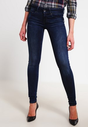 Frau trägt dunkelblaue Skinny-Jeans, ein hochgekrempeltes karierte Hemd und schwarze High Heels, steht vor einem einfarbigen Hintergrund.