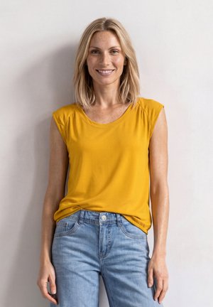 Femme blonde souriante, portant un top jaune sans manches et un jean bleu clair, debout contre un mur blanc uni.