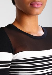 Top a righe bianco e nero con sovrapposizione in nero trasparente, caratterizzato da un collo rotondo e una texture morbida. Motivo a righe orizzontali distintivo.