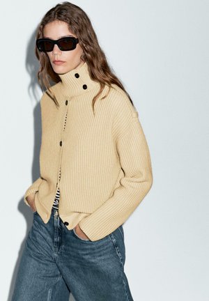 Gelbes geripptes Strickoberteil mit hohem Kragen und drei schwarzen Knöpfen, kombiniert mit blauen hoch taillierten Jeans. Dunkle Sonnenbrillen vervollständigen den Look.