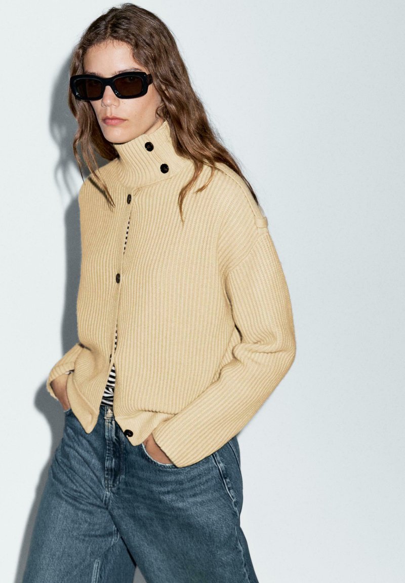 Gul ribbad stickad tröja med hög krage och tre svarta knappar, tillsammans med blå högmidjade denimjeans. Mörka solglasögon fulländar looken.