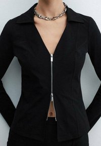 Black zip-up jacket with a V-neckline, long sleeves, and a silver zipper. Features a structured fit and smooth fabric texture.