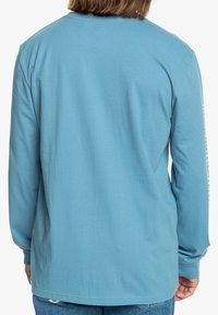 Long-sleeve shirt in light blue cotton, featuring a round neckline and a relaxed fit. Back view shows smooth texture without graphics.
