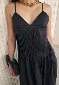 Svart ärmlös klänning med V-ringning, med vertikala veck och en utsvängd kjol. Utsmyckad med ett kraftigt bärnstenshalsband och en vävd handväska.