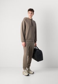 Hanorac maro și pantaloni de trening asortați, realizați dintr-un material moale, purtați cu adidași bej. O geantă neagră este ținută într-o mână.
