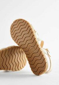 Sandalias blancas con una suela marrón, que presentan un patrón de huella ondulada. Las correas están hechas de un material suave con un acento brillante en la hebilla.