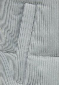 Tela de pana de color gris claro con textura acanalada vertical; presenta un bolsillo tipo parche y costuras visibles. Aspecto suave y color monocromático.