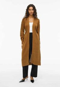 Cardigan long brun, texturé, avec un motif en tricot et une ouverture à l'avant, associé à un haut blanc et un pantalon large noir.