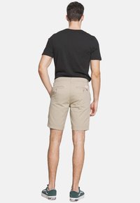 Shorts bej cu două buzunare la spate, croi slim și etichetă pe talie. Purtați cu un tricou negru și adidași verzi, evidențiind un look casual.