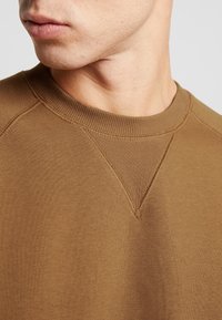 Brun sweatshirt med ribbad halsringning och texturerade axelsömmar. Har en ren, enkel design med subtila söm-detaljer.