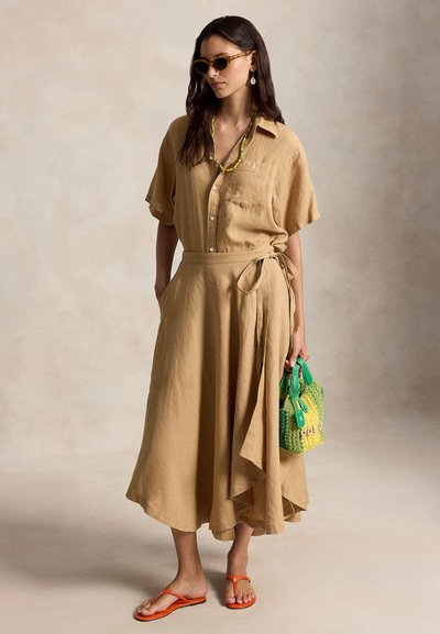 Moteris dėvi smėlio spalvos susegamą suknelę su surišta juosmens juosta, oranžines sandalėles, kiauros spalvos akinius nuo saulės, žalios ir geltonos spalvos rankinę bei karoliukų vėrinį.