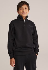 Melnā zipa jakā ar augstu apkakli, ar ribotām piedurknēm un apakšmalu, kā arī subtilu logotipu uz krūtīm. Mīksta auduma tekstūra.