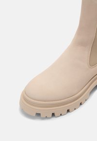 Béžová kotníková bota s hladkým semišovým povrchem, zaoblenou špičkou, elastickými bočními panely a silnou gumovou podrážkou s drážkovaným vzorem.