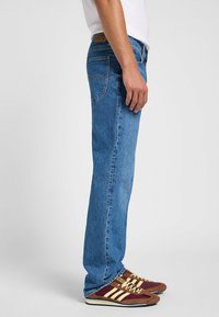 Jeans de mezclilla de color azul claro con corte recto, que presentan un diseño clásico de cinco bolsillos y una etiqueta de marca de cuero marrón en la cintura.