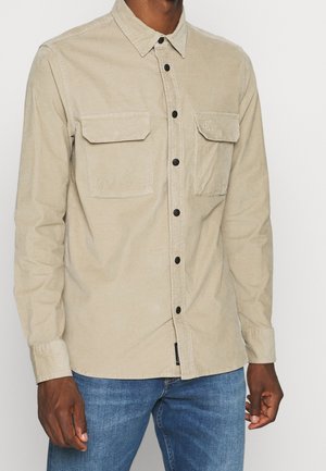 Mand iført lysebeige skjorte med knapper og to brystlommer samt blå jeans, vist fra hals til hofter mod ensfarvet baggrund.