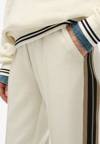 Crème-kleurige joggerbroeken met zijstrips in bruin en zwart. Kenmerken zijn onder andere een geborduurd logo op de heup, geribbelde boorden en een zachte textuur.
