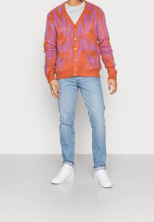 Persoon draagt een oranje-paars gebreid vest over een wit shirt, lichtblauwe spijkerbroek en witte sneakers, staand tegen een witte achtergrond.