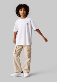 Camiseta de algodón blanca con el logo "CK" en el pecho, combinada con pantalones cargo beige con grandes bolsillos laterales y zapatillas blancas.