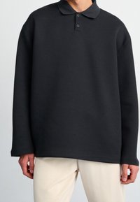 Svart tröja i mjukt material med krage, treknappars knappslå och långa ärmar. Har en avslappnad passform och en minimalistisk design.