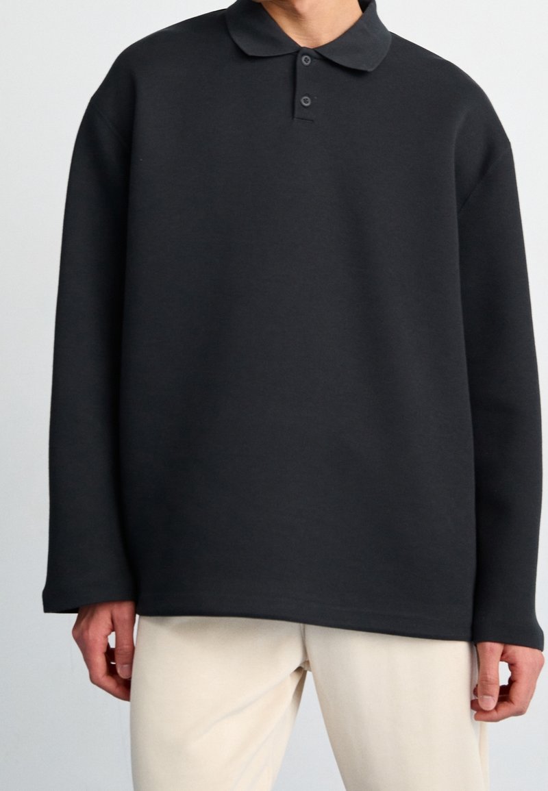 Svart tröja i mjukt material med krage, treknappars knappslå och långa ärmar. Har en avslappnad passform och en minimalistisk design.