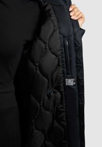 Giacca piumino nera con texture a trapuntatura, tasca con zip e etichetta logo all'interno. Finitura in satin liscia, progettata per il calore.