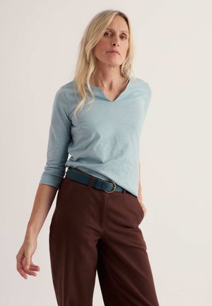 Femme aux longs cheveux blonds portant une chemise bleu clair à manches longues, un pantalon marron et une ceinture bleue, debout devant un fond clair uni.
