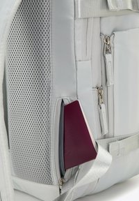Mochila plateada y gris con un bolsillo de malla, múltiples cremalleras y un objeto morado parcialmente visible en un compartimento lateral.