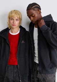 Black zip-up hoodies with front pockets, worn over a red shirt with vertical stripes and a white tank top. Light and dark textured fabrics.