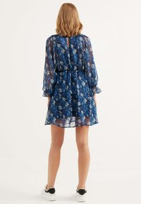 Blauwe bloemen mini-jurk van doorschijnende stof, met lange pofmouwen, een sleutelgat achterkant en een elastische taille. Kenmerkt zich door witte en lichtblauwe bloempatronen.