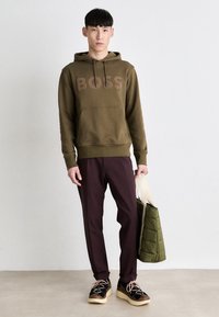 Olivegrüner Hoodie mit einem "BOSS"-Logo, kombiniert mit kastanienbraunen Hosen und schwarzen Sneakers. Hält eine grüne, gesteppten Tragetasche.