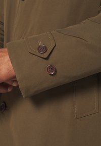 Manche de manteau marron avec deux boutons marron foncé et coutures visibles, portée par une personne à la carnation moyenne.