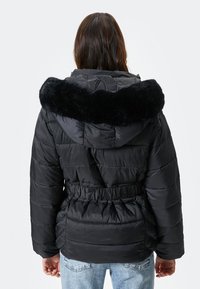 Schwarze Daunenjacke mit großem Kunstfellkragen, elastischer Taille und gestepptem Muster. Die Rückseite hat eine glatte Verarbeitung und Raffung.
