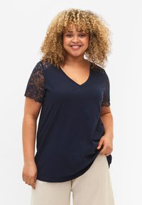 Camiseta navy de cuello en V con mangas cortas que presenta intrincados detalles de encaje negro en los hombros. La tela suave cae suavemente alrededor del cuerpo.